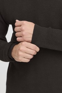 Dark textured pullover with a waffle weave design, featuring ribbed cuffs. Two hands grasp the sleeve, highlighting material and fit.