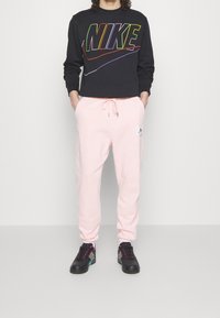 Svart sweatshirt med flerfärgad "NIKE"-logotyp, ihopparad med ljusrosa joggingbyxor som har en logotypapplikation och resåren i midjan. Svarta sneakers.