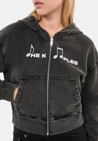 Donkergrijze capuchontrui met ritssluiting en voorklepzak, met witte muzieknoten en de tekst "THE K PLES." De stof heeft een verweerde, gestructureerde uitstraling.