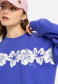Sudadera morada con un estampado floral blanco de flores de hibisco en el pecho. Cuello y puños de canalé, corte casual.