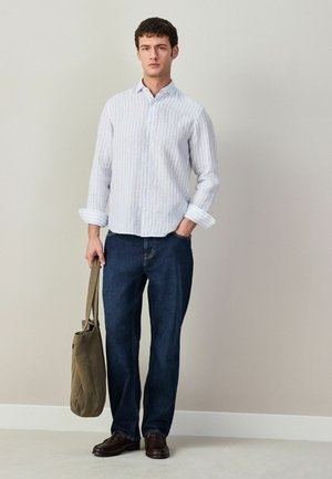 Junger Mann trägt ein hell gestreiftes Hemd mit Knöpfen, dunkle Jeans, braune Schuhe und hält eine olivgrüne Tragetasche vor einer schlichten beigen Wand.