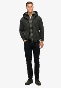 Felpa verde scuro con cappuccio e cordini sopra una camicia a quadri; jeans neri aderenti; scarpe nere con dettagli testurizzati; figura maschile in piedi.