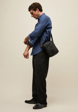 Vyras prisitaiko džinsinio marškinio rankoves, dėvi juodas kelnes ir juodą per petį nešiojamą rankinę ant šviesaus vientiso fono.