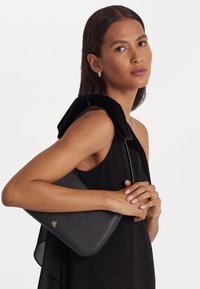 Mujer con cabello largo y oscuro que lleva un vestido negro sin mangas sostiene un bolso texturizado negro con el logo dorado de RLL en su hombro.