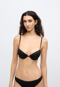 Γυναίκα με μακριά σκουρόχρωμα μαλλιά, φορώντας μαύρο σουτιέν και ταιριαστό μαύρο εσώρουχο, στέκεται μπροστά σε απλό ανοιχτόχρωμο φόντο.