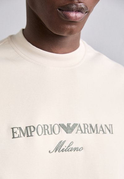 Crème kleurig katoenen T-shirt met een ronde hals en korte mouwen; voorzien van geborduurd "EMPORIO ARMANI Milano" logo in lichtgroen.