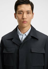 Chaqueta azul marino oscura con grandes bolsillos frontales, cuello afilado y textura suave, superpuesta sobre una camisa de patrón azul claro.