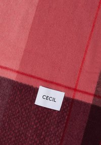 Tissu à carreaux rouge et bordeaux avec une étiquette rectangulaire blanche portant l'inscription "CECIL" cousue sur la surface.