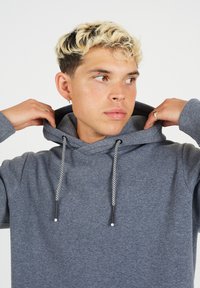 Felpa grigia con cappuccio, dotata di lacci intrecciati, texture morbida e vestibilità rilassata. Il modello sistema il cappuccio, mettendo in evidenza il design.