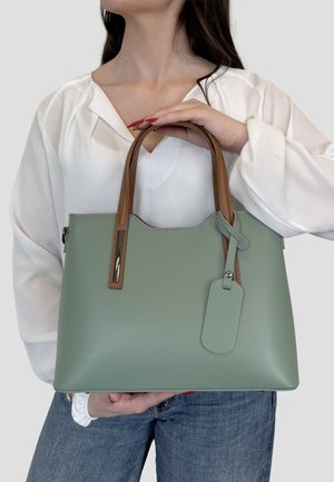 Kobieta w białej bluzce i dżinsach trzyma zieloną torebkę z brązowymi uchwytami i dopasowaną zawieszką na jednolitym tle.