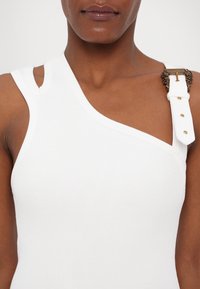 Gros plan d'une personne portant un haut asymétrique sans manches blanc avec une sangle décorative à boucle dorée sur l'épaule droite.