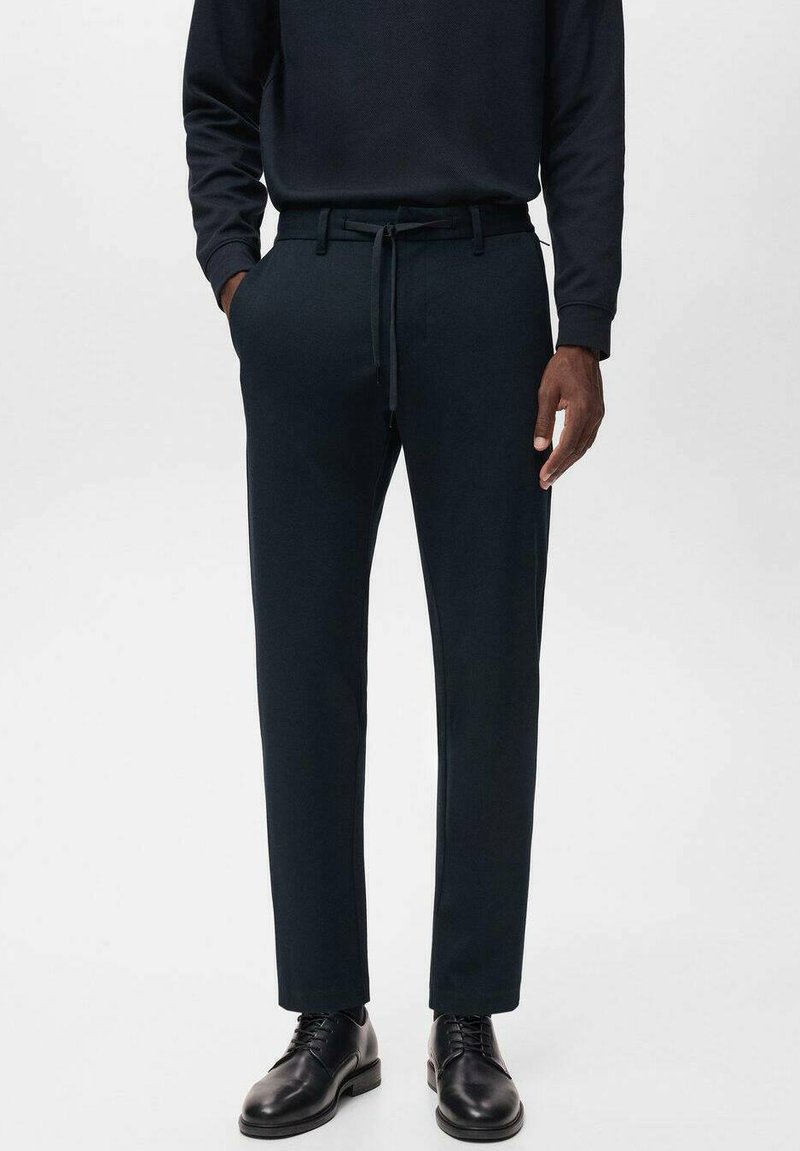 Pantalons bleu marine foncé en tissu doux, avec une taille à cordon de serrage, des poches latérales et une coupe ajustée, associés à des chaussures noires brillantes.