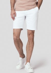 Białe bawełniane szorty z płaskim przodem i bocznymi kieszeniami, noszone z białymi trampkami. Materiał wydaje się gładki i ma luźny krój.