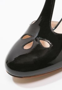 Bout de chaussure en cuir verni noir avec trois découpes en forme de goutte et doublure intérieure beige visible à travers les ouvertures.