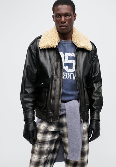 Veste bomber en cuir noir avec un col en fausse fourrure beige, fermeture éclair à l'avant et poches latérales ; portée sur un sweat gris et un pantalon à carreaux.