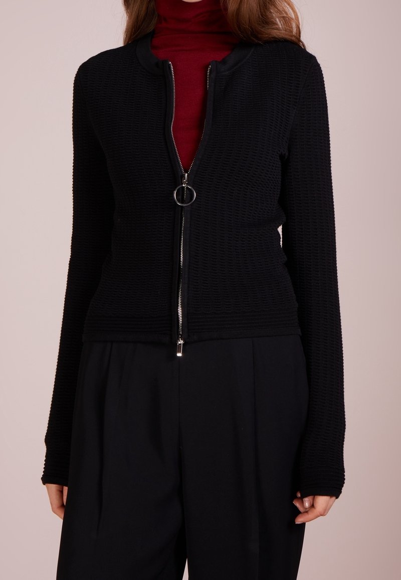Femme portant une veste noire texturée avec fermeture éclair à anneau sur un col roulé rouge et un pantalon noir à jambes larges.