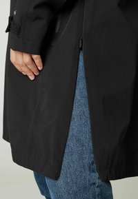 Svart, vattentålig jacka med dragkedja och kardborredetaljer vid ärmarna, visad tillsammans med blå denimjeans. Har en sidoklyvning vid fållen.