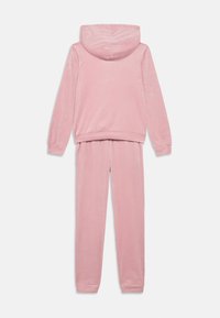 Rožinės spalvos laisvalaikio kostiumas su gaubtu, pagamintas iš minkštos medžiagos; turi ilgomis rankovėmis, elastines rankogalius ir laisvo silueto kelnes su elastiniu juosmeniu.