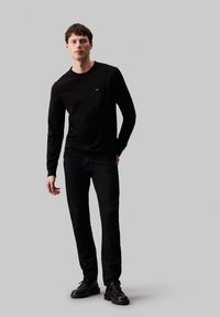 Svart langermet T-skjorte med ribbestrikket rund hals, kombinert med svarte jeans og svarte snørestøvler, med en liten logo på brystet.