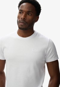 Biała bawełniana koszulka z okrągłym dekoltem i krótkimi rękawami. Posiada subtelne, tonowe logo na piersi. Gładka tekstura, luźny krój.