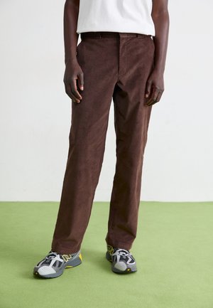 Brown corduroy pants with a straight cut, visible texture, and a belt loop. Paired with gray and yellow athletic shoes.