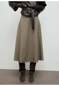 Une personne portant une veste en cuir noir à ceinture, une jupe midi beige à carreaux et des bottes en daim marron décontractées jusqu'aux genoux, se tenant sur un sol gris.