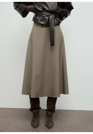 Une personne portant une veste en cuir noir à ceinture, une jupe midi beige à carreaux et des bottes en daim marron décontractées jusqu'aux genoux, se tenant sur un sol gris.