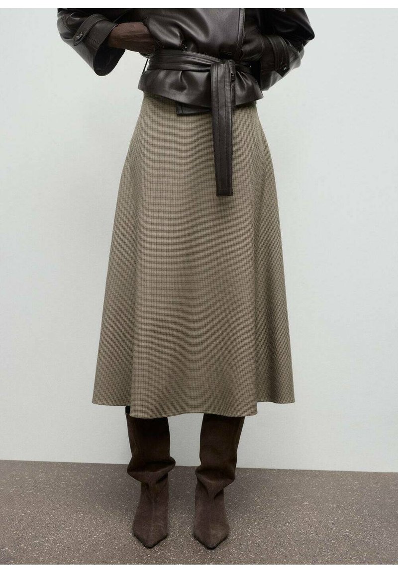Une personne portant une veste en cuir noir à ceinture, une jupe midi beige à carreaux et des bottes en daim marron décontractées jusqu'aux genoux, se tenant sur un sol gris.