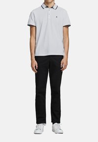 Polo shirt blanc avec des accents de col noir, associé à un pantalon noir et des baskets blanches. La chemise arbore un logo sur la poitrine.