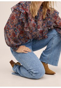 Femme à genoux portant un chemisier transparent à fleurs avec volants, un jean large bleu clair et des bottines en suède beige à talons carrés.