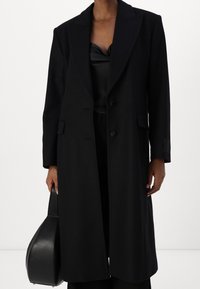Sort uldblandingsfrakke med en lang, lige silhuet, slagkraver, to knapper og sidelommer. Samlet med en slank sort top og taske.