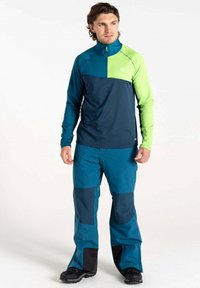 Blaue und grüne Langarmbluse mit halbem Reißverschluss und kontrastierenden Einsätzen, kombiniert mit blauen Hosen mit dunkleren Akzenten und strapazierfähigem Stoff.