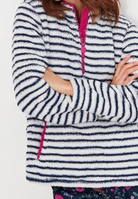 Osoba ubrana w puszysty biały i granatowo paskowany sweter z różowym zamkiem, w kwieciste spodnie, z założonymi rękami.