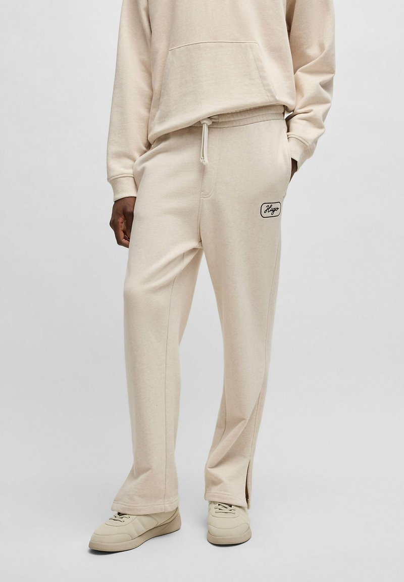 Beige Baumwollmischung Jogginghosen mit einem Kordelzugbund, seitlichen Taschen und einem schwarzen Logopatch auf der Rückseite. Die Hose hat eine lockere Passform und gespaltene Säume.