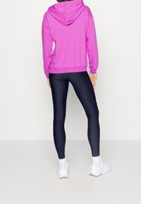 Felpa rosa con cappuccio e vestibilità comoda, abbinata a leggings blu scuro e sneakers bianche. Il tessuto appare morbido con una texture liscia.