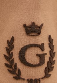 Tessuto a maglia marrone con una lettera "G" ricamata di un marrone scuro, una corona sopra e foglie di alloro ai lati, con dettagli testurizzati.