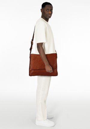 Bolso mensajero de cuero marrón con textura lisa, con cierre de solapa y una correa ajustable, sostenido en una postura de pie.