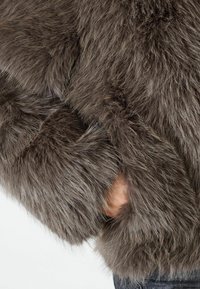 Hnedá umelá kožušina bunda s hrubým, textúrovaným povrchom, obsahujúca skrytú kapsu a voľný, oversized strih. Mäkka a nadýchaná vzhľad.
