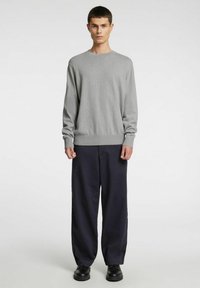 Hall hall kootud kampsun pika varrukaga, ümara kaelusega ja soonikkoes äärega, mida on kombineeritud lahtiste tumesiniste pükstega ja mustade kingadega.