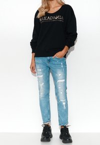 Černá mikina s nápisem "MAKADAMIA" v lesklých písmenech, v kombinaci se sepranými světle modrými džíny a černými teniskami. Casual střih a uvolněný styl.