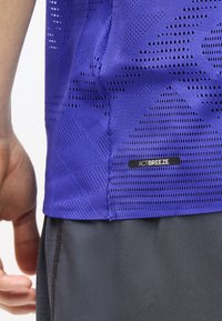 Κοντινό πλάνο ενός ατόμου που φοράει μωβ διαπνέον μπλουζάκι από δίχτυ με την ένδειξη "ACTIBREEZE" και αθλητικές σκούρες γκρι βερμούδες.