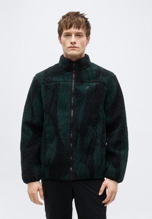 Νέος άνδρας που φοράει ζακέτα φλις με φερμουάρ σε σκούρο πράσινο και μαύρο μοτίβο, στέκεται μπροστά σε απλή φωτεινή επιφάνεια.