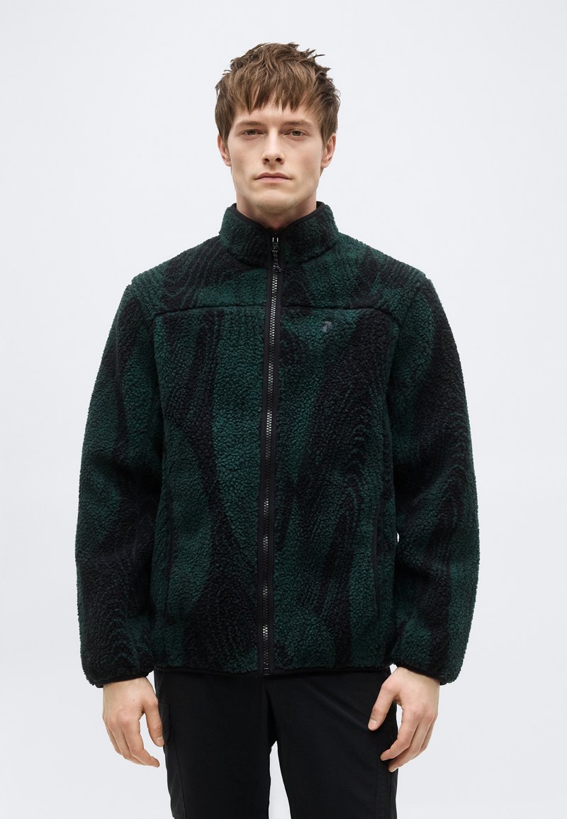 Νέος άνδρας που φοράει ζακέτα φλις με φερμουάρ σε σκούρο πράσινο και μαύρο μοτίβο, στέκεται μπροστά σε απλή φωτεινή επιφάνεια.