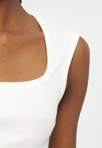 Κοντινό πλάνο ενός ατόμου που φοράει λευκό αμάνικο ριμπ τοπ με τετράγωνη λαιμόκοψη σε απλό φόντο.