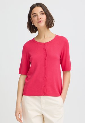 Femme portant un cardigan à manches courtes boutonné rose vif et un pantalon beige clair, debout devant un fond blanc uni.