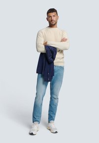 Beige gebreide trui met ronde hals, blauwe jeans en witte sneakers. Houdt een donkerblauwe jas vast, staat met gekruiste armen tegen een lichte achtergrond.