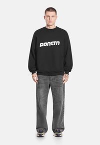 Czarna bluza z białym, pogrubionym napisem "PRNSTN" na piersi, w zestawie z szarymi jeansami i czarnymi sneakersami, o luźnym kroju.
