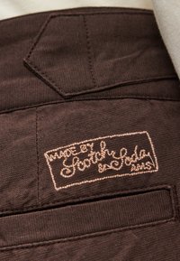 Καφέ ύφασμα με υφή στην επιφάνεια που διαθέτει τσέπη με πατς και κεντημένη ετικέτα "Made by Scotch & Soda" σε ανοιχτό μπεζ χρώμα.