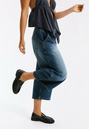 Person iført løs mørkeblå ærmeløs top, brede korte blå jeans og sorte læder loafers, poserende med det ene ben løftet.