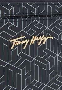 Navy portemonnee met geometrisch wit patroon, met een gouden "Tommy Hilfiger" logo in cursieve letters. Textuurleer afwerking, compact ontwerp.